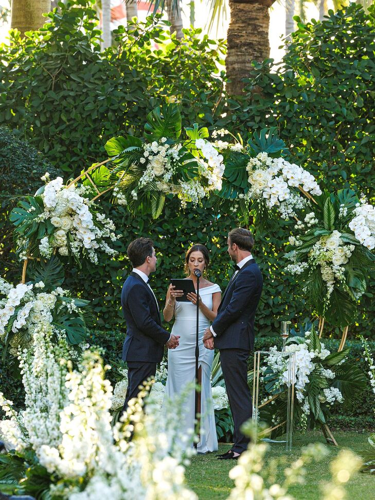 Grooms in Deep Blue Tuxedos At Lush, Tropical Altar With White Flowers With Officiant
