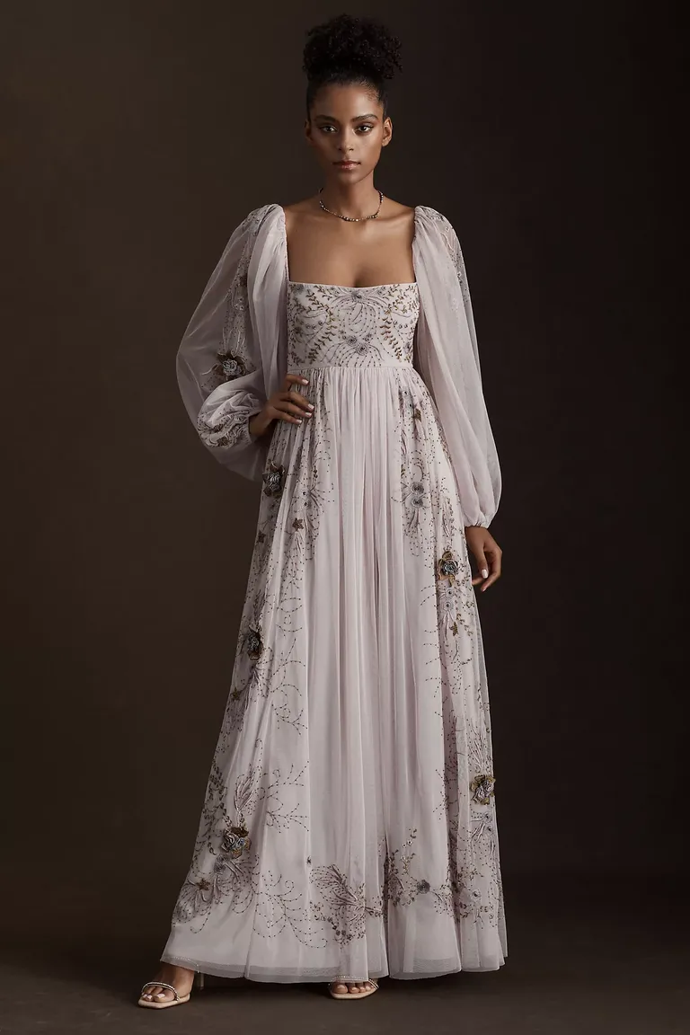 Model wears a lavender colored floor-length dress with long sleeves and floral beading. 