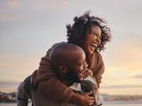 young couple laughing on the beach while man carries woman piggyback 