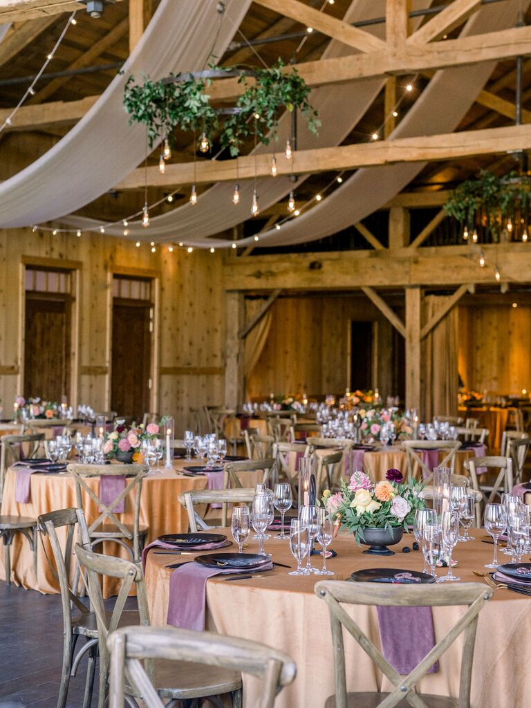 rustic wedding reception venue with draped ceilings, greenery chandeliers, string lights, blush tablecloths, purple cloth napkins, and wooden cross back chairs