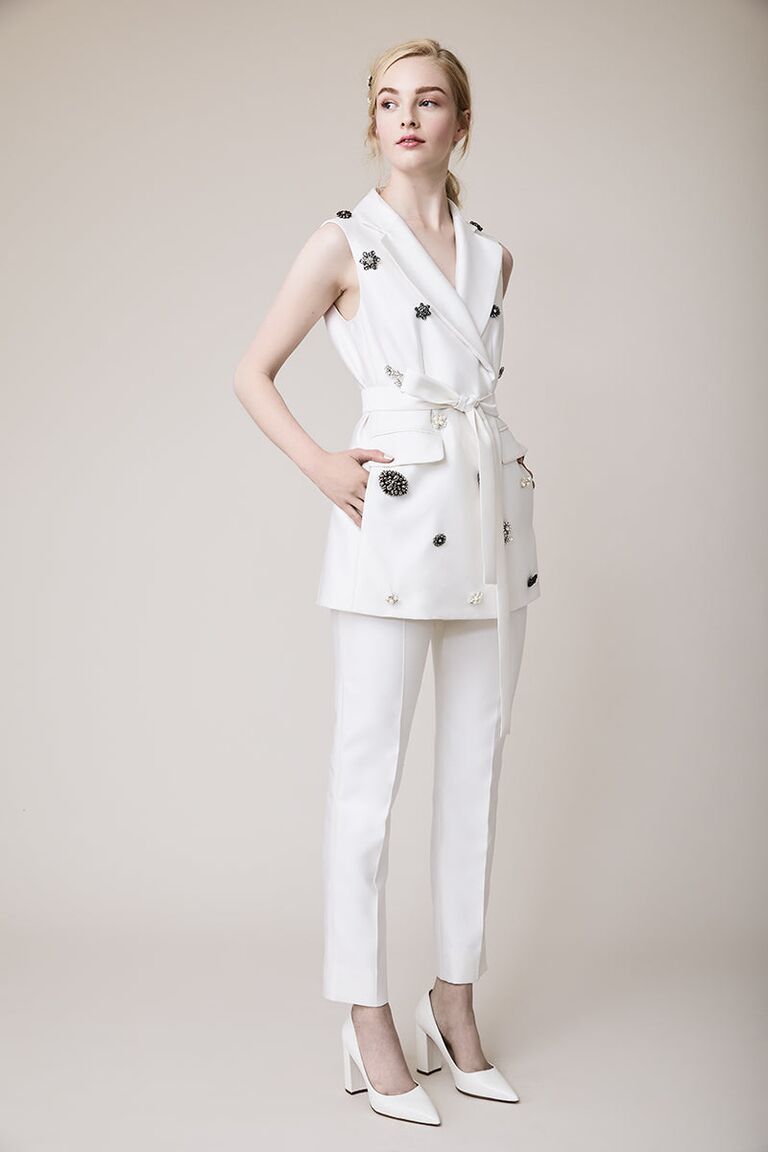Model wears white fitted trousers and a belted vest with floral embellishments. 