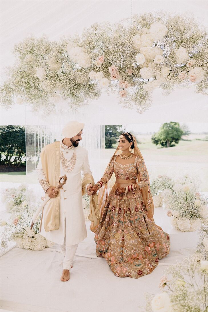 A glamorous Sikh bride and groom in a traditional lengha (in gold, red and green) and achkin (in white) with a turban and a sword at their Guru Granth Sahib altar after their luxurious religious ceremony at a golf course venue.