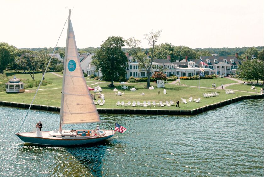 A sailboat glides through the water, with a charming inn visible on the shore in the background.