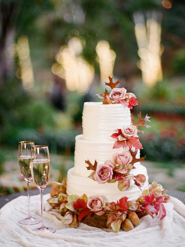 elegant fall wedding cake with white buttercream icing, red fall leaves and mauve roses next to two champagne glasses