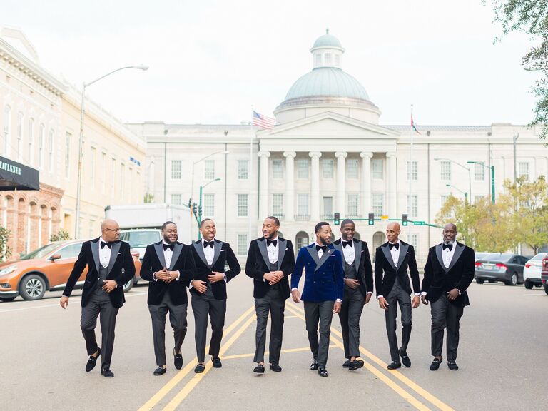 groom wearing navy blue velvet tuxedo walks down the street next to groomsmen in black tuxedos